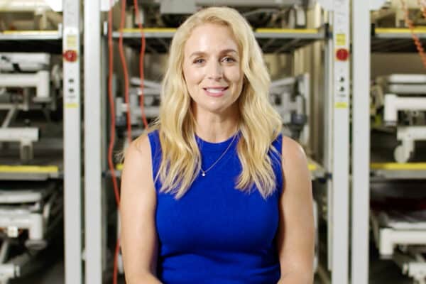 woman smiling into camera, waist up, blue dress, blonde hair, hospital beds stacked behind her