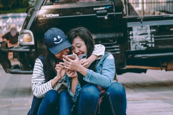 Two girls laughing and smiling on the sidewalk while looking at their phones